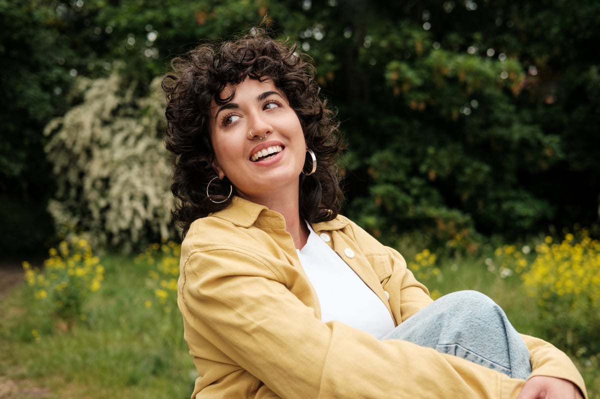 Cheerful young woman with curly hair, wearing a vibrant yellow jacket and jeans, sitting in a serene park, smiling joyfully